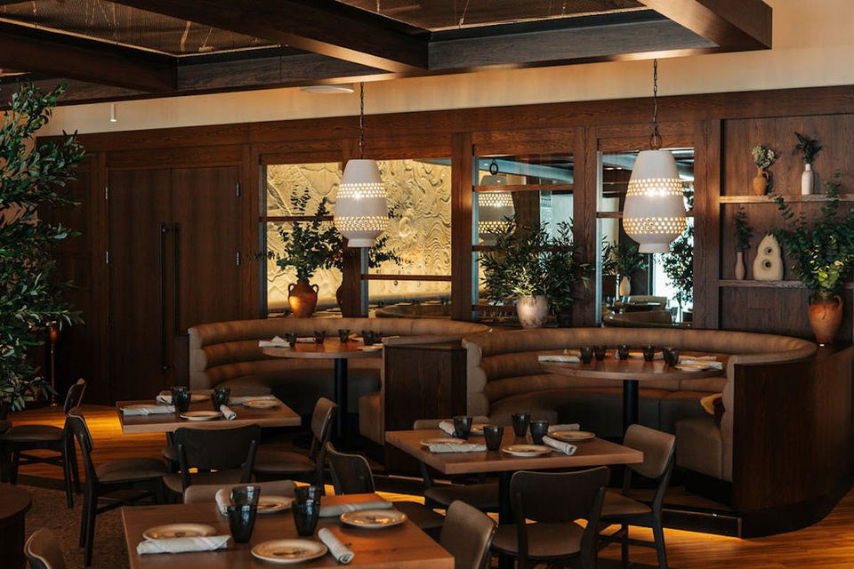 The interior of Naia Chicago restaurant features a mix of wooden tables and chairs, along with curved booth seating. Two large, white, perforated