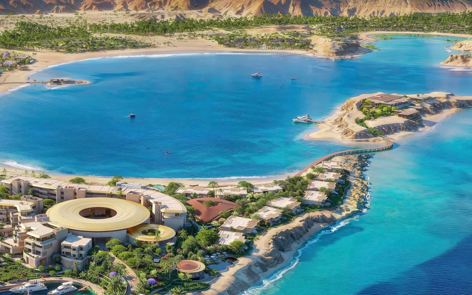 Aerial view of a luxury coastal resort development with modern circular architecture, beachfront villas, and turquoise waters backed by desert mountai