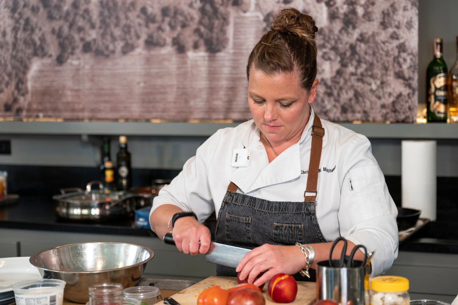 Chef Christina Machamer, with her hair in a bun, wearing a white chef's coat and a denim apron, chops fruit on a cutting board.