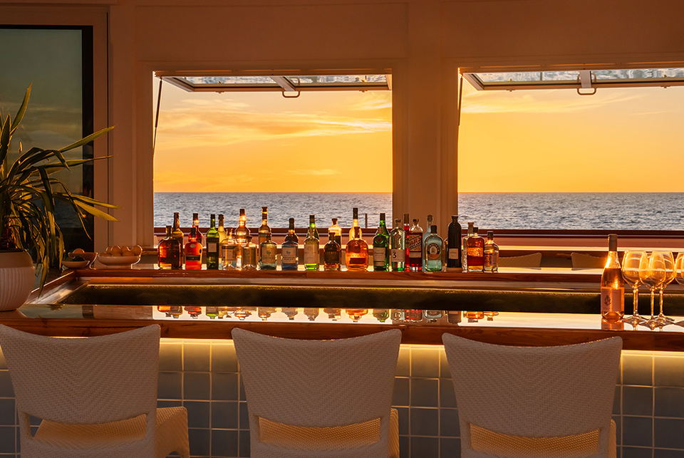 A high-end bar interior with ocean sunset view through large windows, matching the documented 'Sunset Bar' feature from Naples Beach Club; Forbes Trav