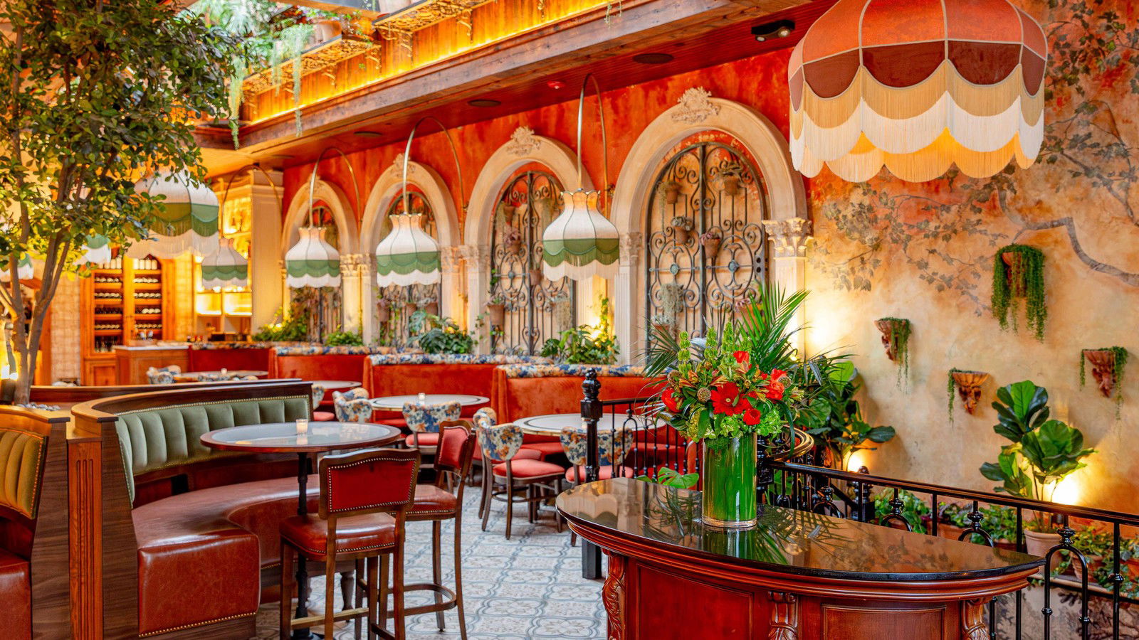A richly decorated restaurant interior with ornate wrought-iron gates, botanical murals, patterned floor tiles, velvet banquettes, and pendant lamps.