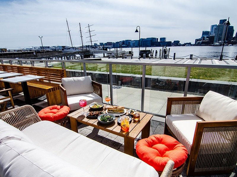 An outdoor waterfront terrace with wooden furniture, white cushions, orange accent pillows, food and drinks on a coffee table, and the Boston city