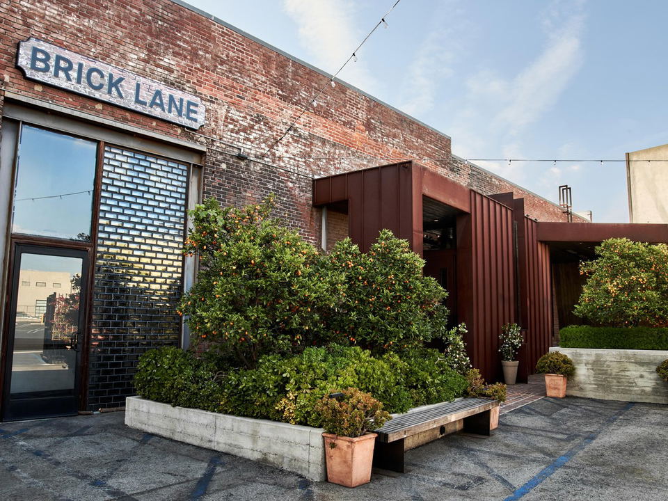 Exterior of Brick Lane restaurant in the Arts District, featuring a brick facade with a 'BRICK LANE' sign, lush trees with orange fruits, a wooden