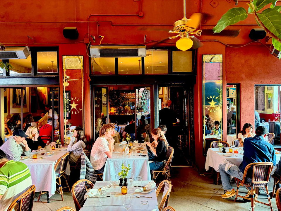 The outdoor dining area of Cafe Stella in Silver Lake, featuring white-clothed tables, rattan chairs, and a bustling lunch crowd under warm red walls