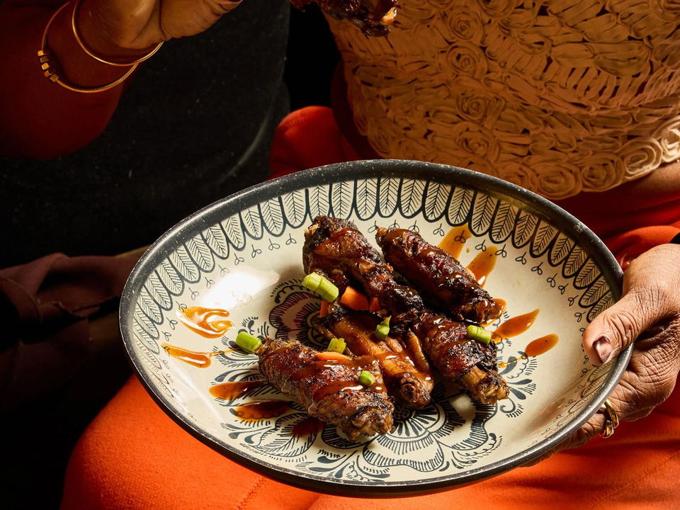 A person in an orange outfit holds a decorative plate of glazed, sauced egg rolls garnished with scallions, with one piece held up in the other hand.