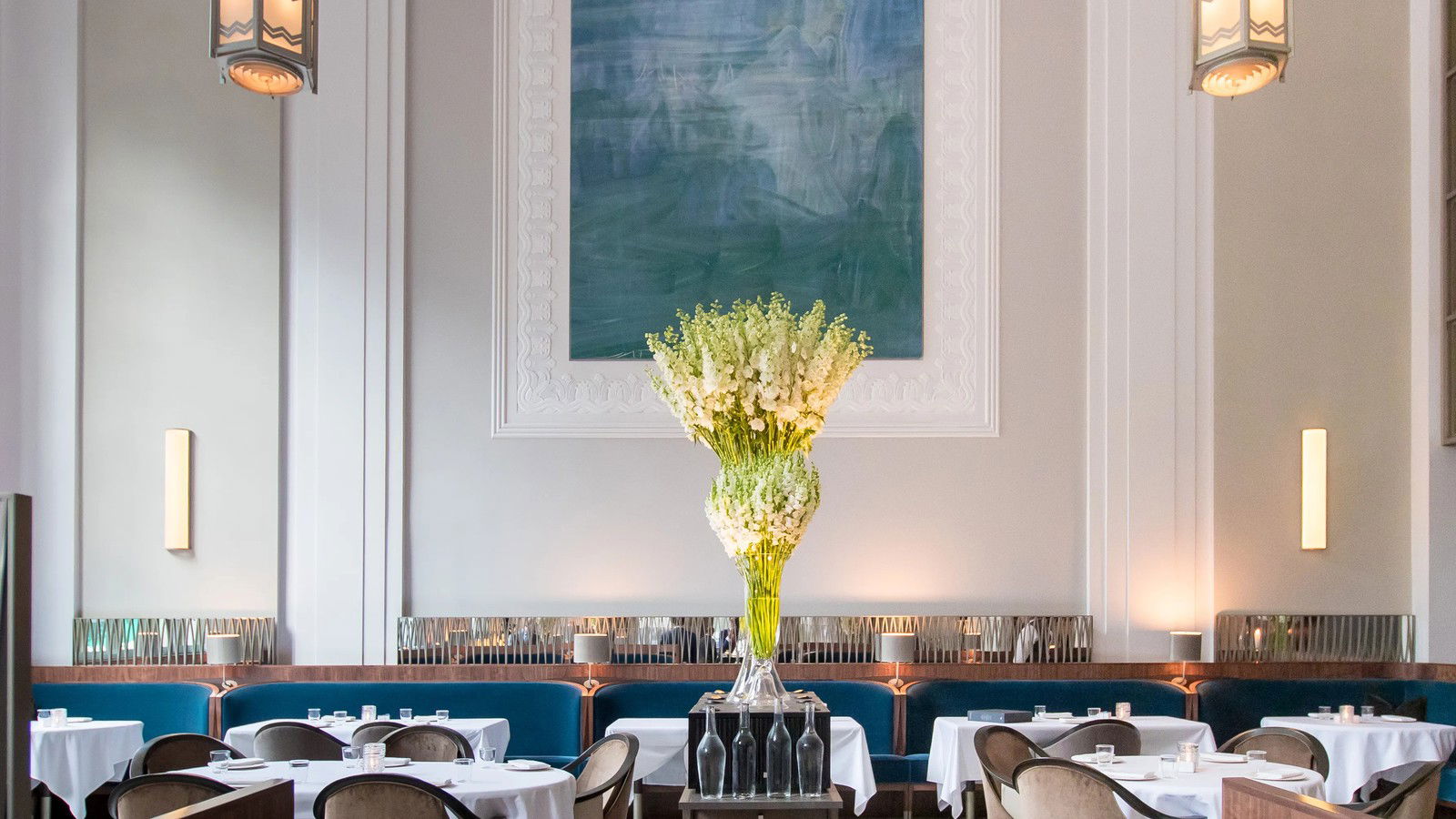 An elegant dining room with high ceilings, teal banquettes, white tablecloths, and a large abstract blue painting.