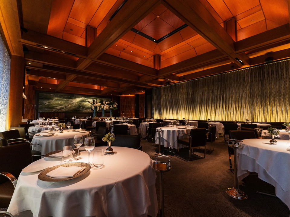 An elegant, warmly lit fine-dining room with white-tablecloth round tables, wine glasses, upholstered chairs, and a large seascape painting.