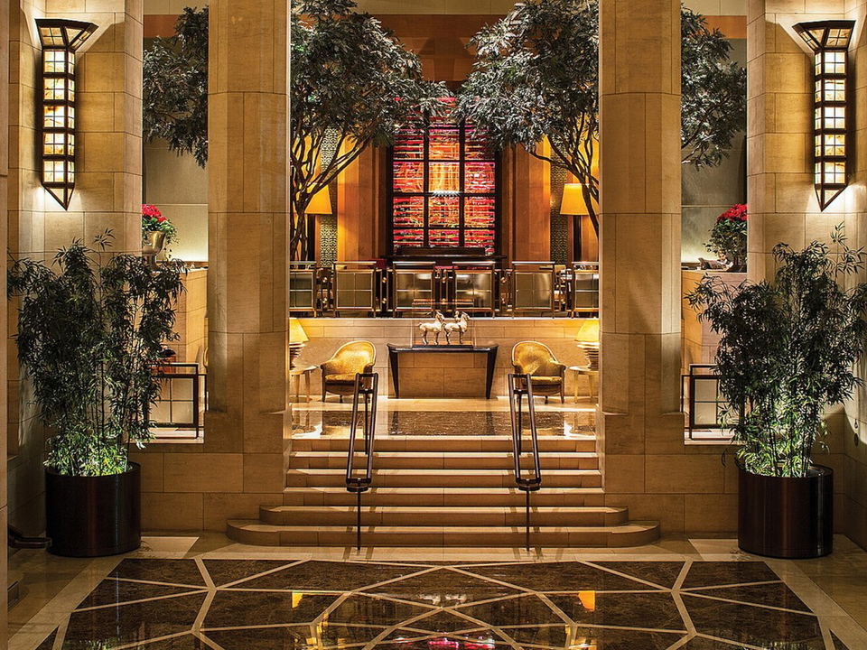 A grand, warmly lit luxury hotel lobby with marble floors, tall stone columns, indoor trees, and a raised seating area.