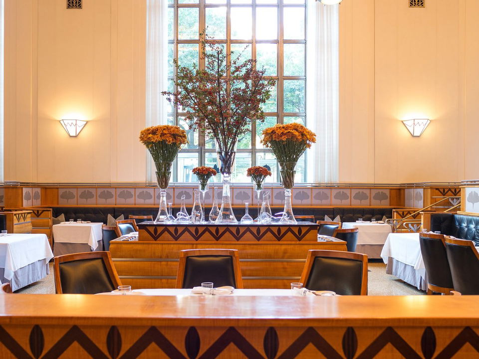 An elegant, high-ceilinged dining room with Art Deco-influenced woodwork, tall multi-pane windows, leather banquettes, white-tablecloth tables, and