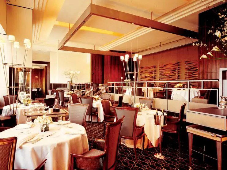 An elegant, warmly lit fine-dining room with white-tablecloth tables, upholstered chairs, and rich wood paneling.