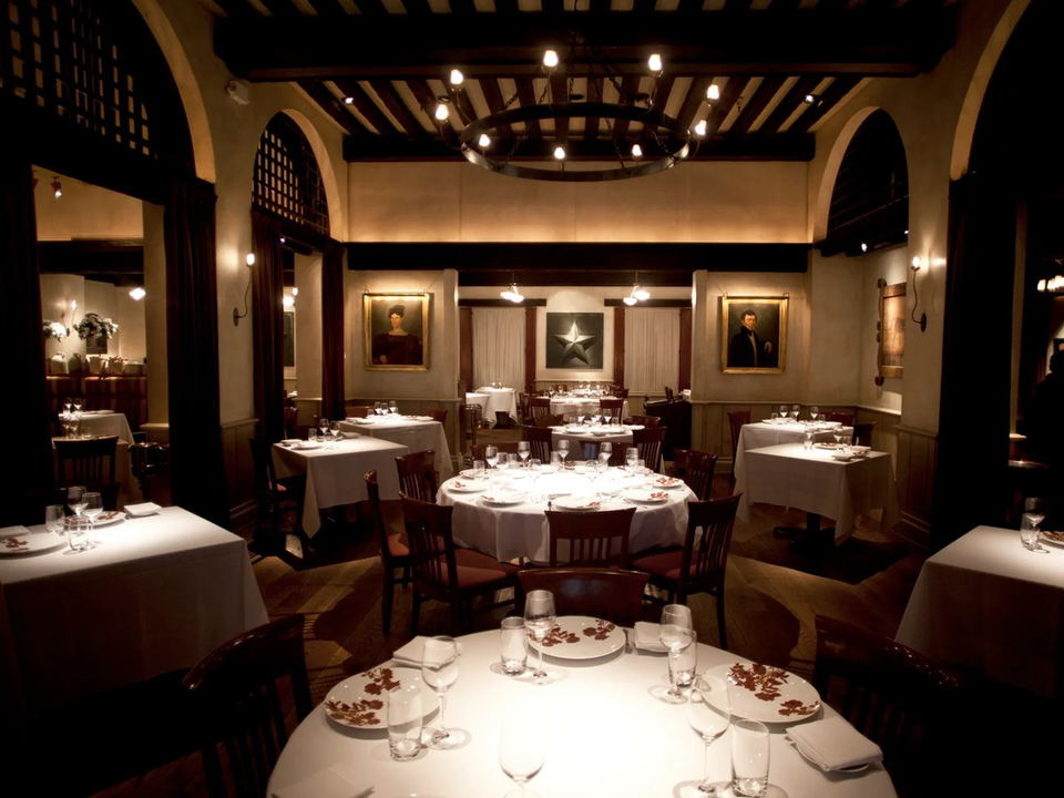 An elegantly set, warmly lit fine-dining room with white-tablecloth round tables, dark wood chairs, arched architectural details, portrait paintings