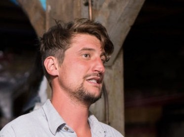A man with a beard and light blue shirt gestures while speaking in a dimly lit stone cellar with oak barrels.