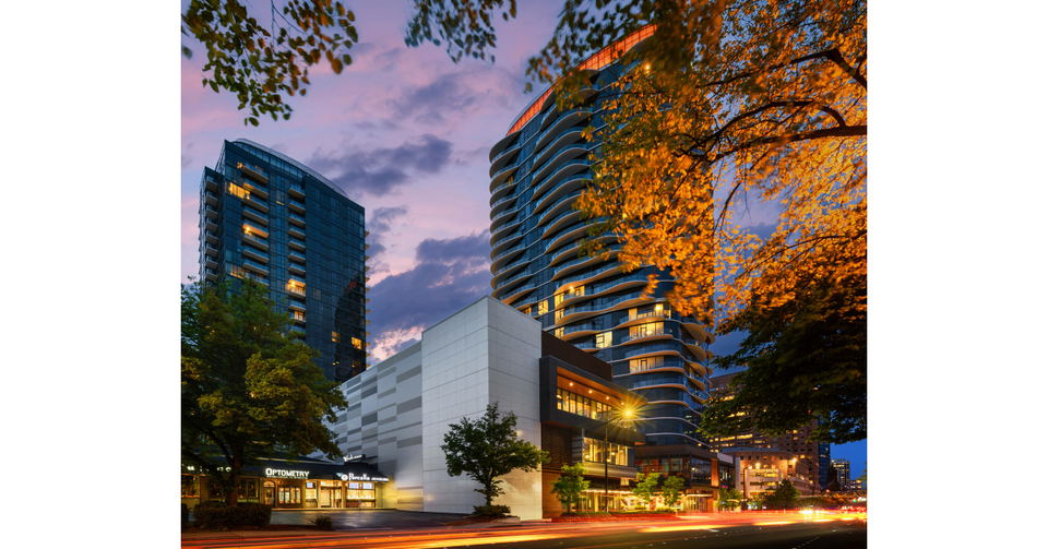 Two modern high-rise buildings with balconies and illuminated windows, framing a purple and pink sunset sky, with autumn trees.