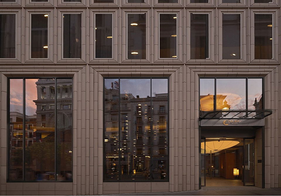 Modern hotel entrance at dusk with glass doors and 'NOBU' signage visible above the entrance; the source page explicitly attests this is 'Entrada al n