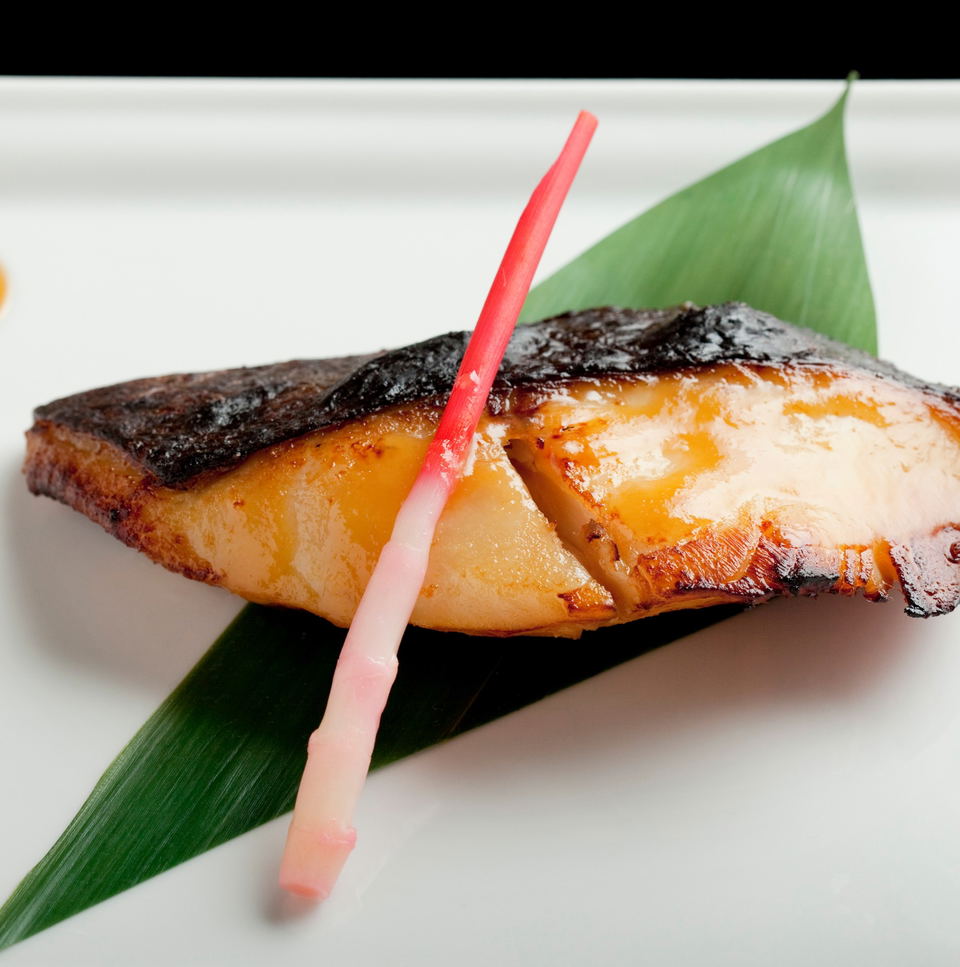 A piece of glazed miso black cod with charred skin on a green leaf, presented in Japanese style; source page explicitly identifies this as 'Miso black