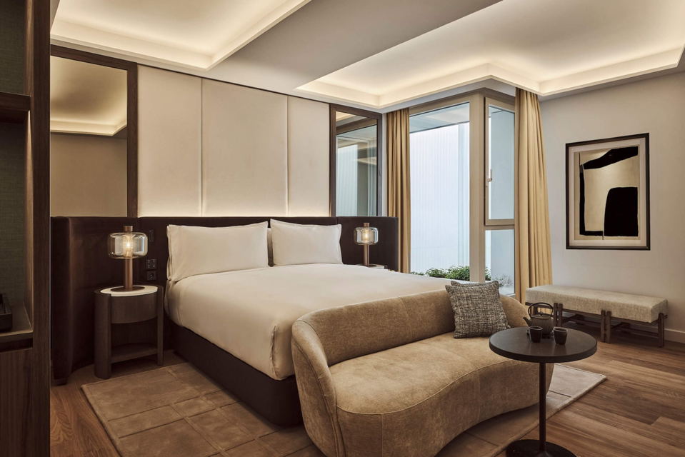 A luxury hotel suite bedroom with warm wood flooring, floor-to-ceiling windows, contemporary furnishings including a beige sofa, and modern lighting;