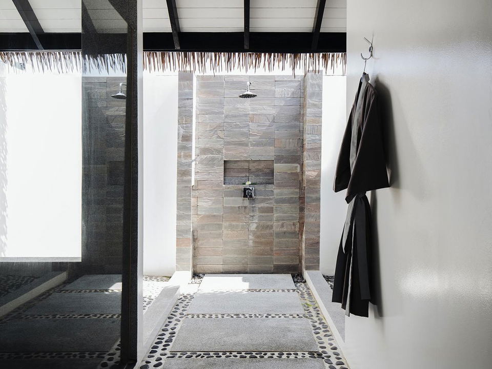 An open-air outdoor shower area at Noku Maldives, featuring natural stone tile walls, pebble flooring, rain showerheads, and dark robes hanging on a
