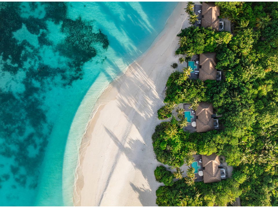 Aerial view of a pristine white-sand beach with turquoise water, coral reefs, lush green vegetation, and thatched-roof villas with private pools.
