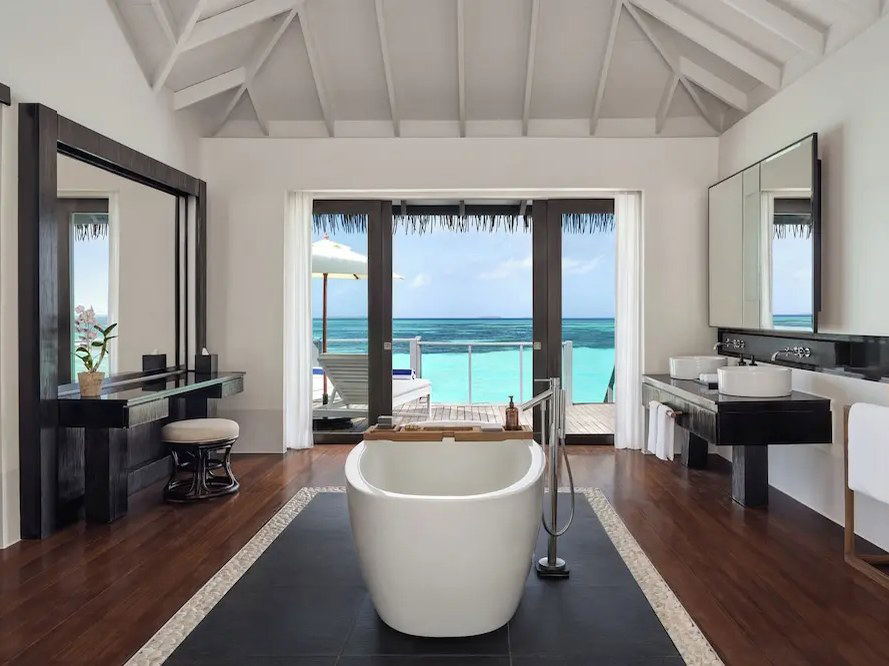 A luxurious overwater villa bathroom at Noku Maldives, with a freestanding tub, dark wood floors, and glass doors opening to the turquoise lagoon.