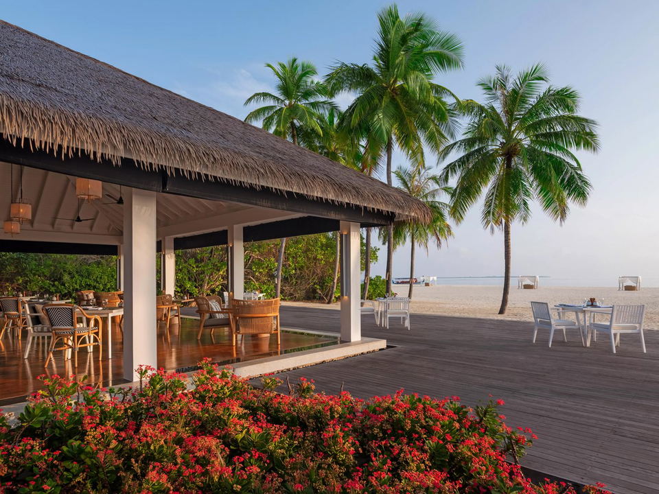 An open-air restaurant with a thatched roof and wooden deck, featuring dining tables with chairs, next to a white sand beach with palm trees.