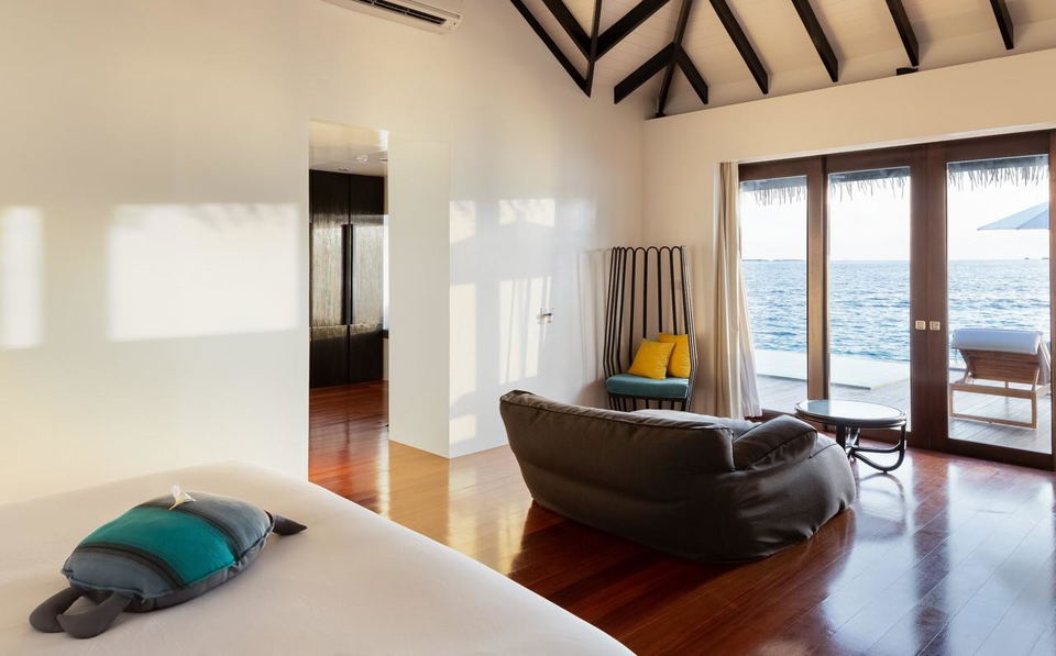 A bright bedroom in Noku Maldives with a bed, a beanbag chair, and a view of the ocean from large glass doors.