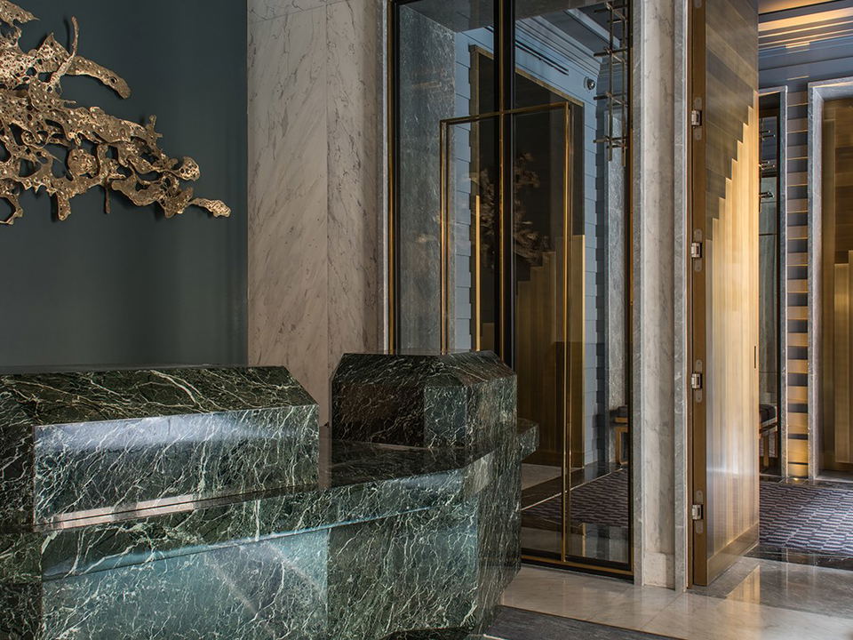 A luxurious hotel lobby with a green marble reception desk, white marble walls, brass-framed glass doors, and a gold organic wall sculpture.