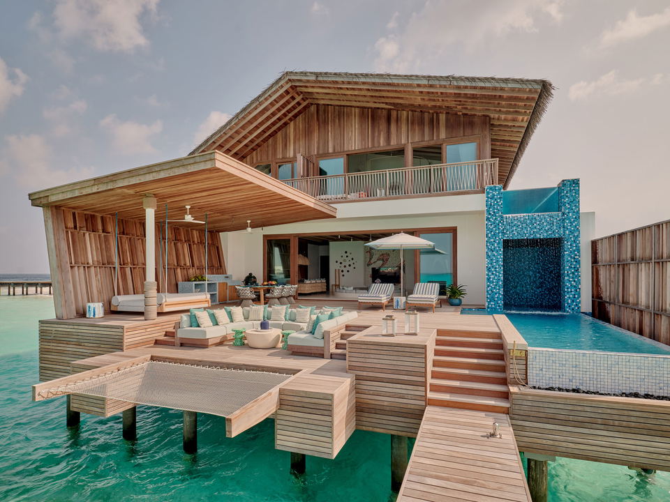 Wide establishing shot of a multi-level overwater villa with thatched roofs extending over turquoise lagoon water, featuring terraced outdoor spaces, 