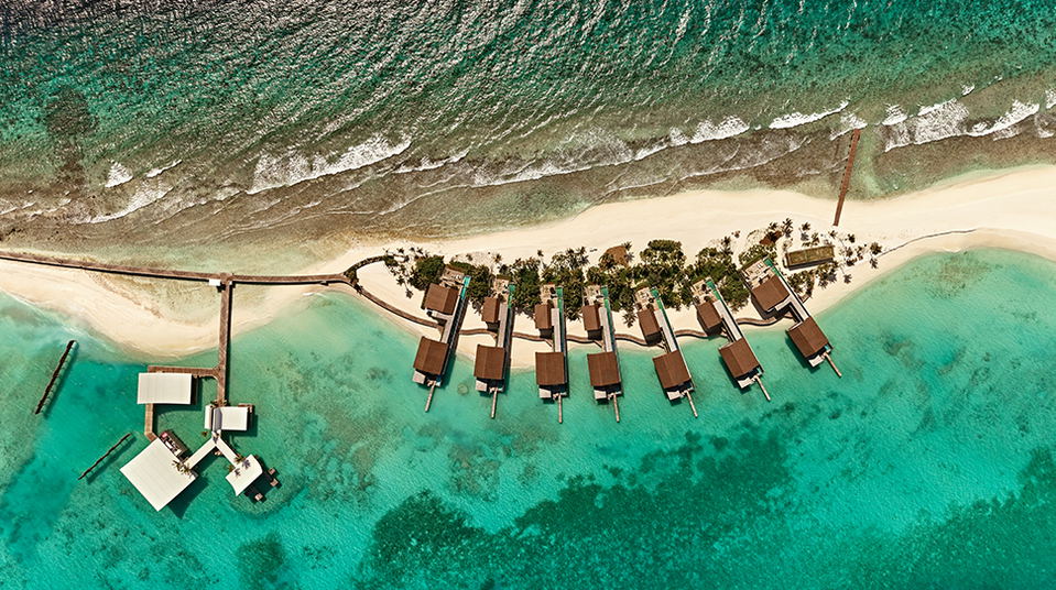 Overhead shot of a tropical resort with brown wooden bungalows on stilts in shallow turquoise water, connected by wooden walkways to a sandy beach lined with palm trees.