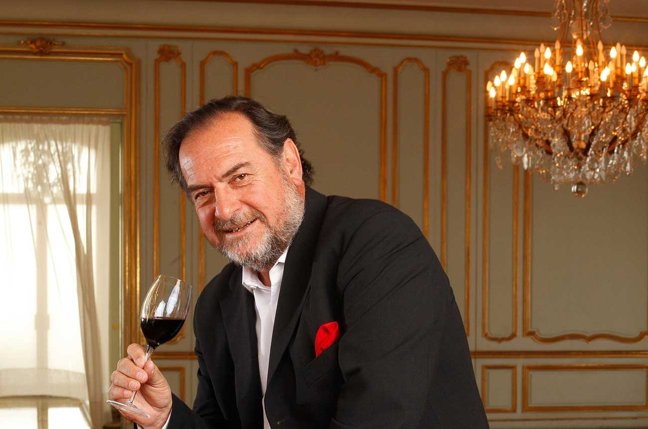A professional portrait of a man in formal attire holding a wine glass in an elegant room with chandelier and ornate wall paneling; the source page fr