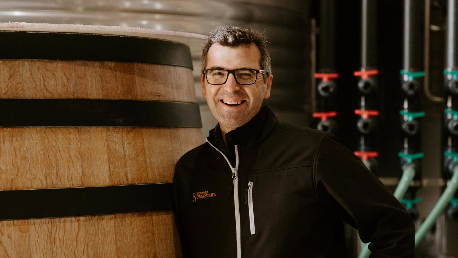 A man in glasses and a black Bodega Numanthia vest poses beside a large wooden aging barrel in a cellar; the source page caption explicitly identifies