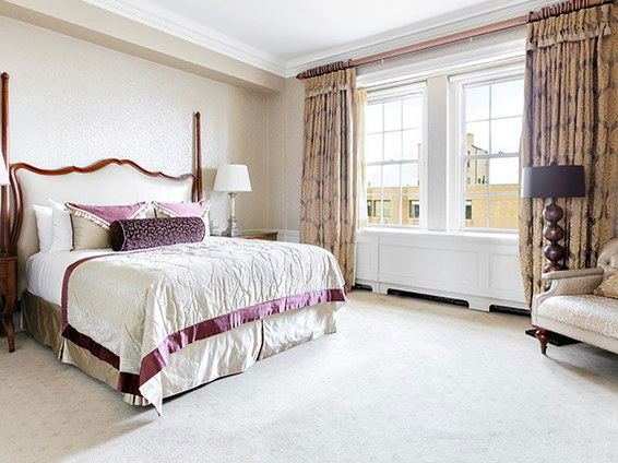 An elegant bedroom at The Pierre, A Taj Hotel, featuring a four-poster bed with cream and purple bedding, and windows overlooking New York City.