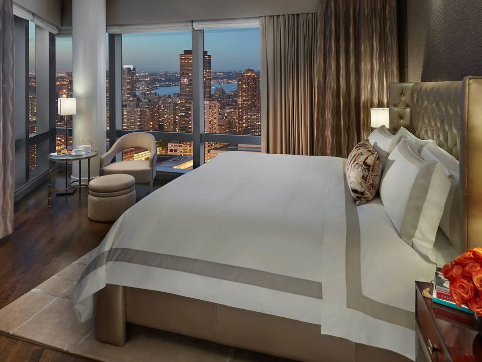 A luxury hotel bedroom at Mandarin Oriental, New York, with a king bed, tufted headboard, and floor-to-ceiling windows overlooking a city skyline at