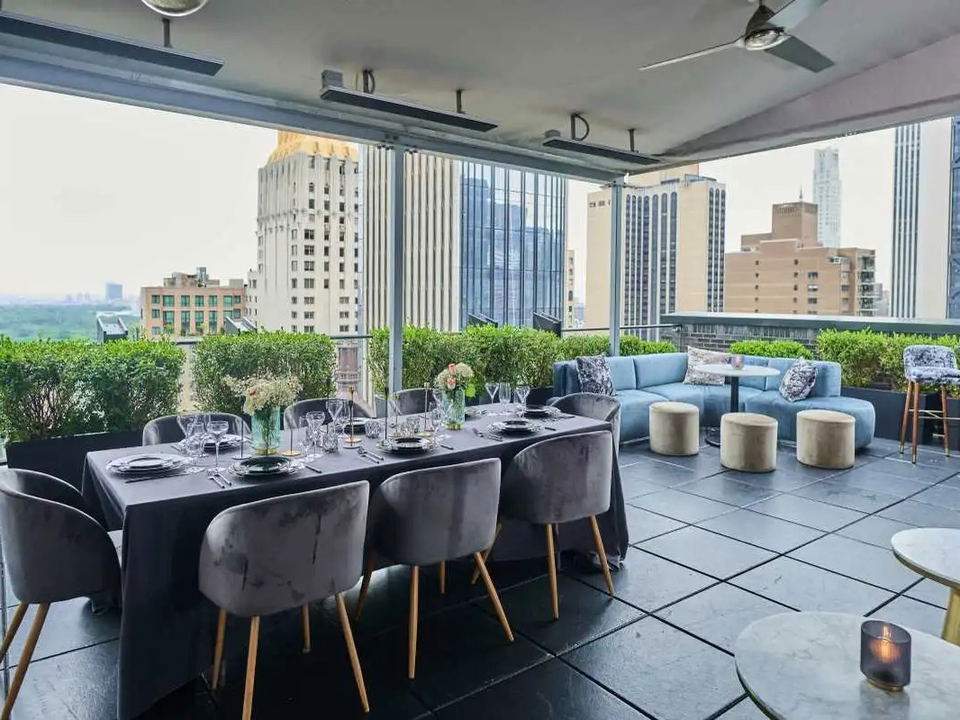 A stylish rooftop terrace with a long dining table, velvet chairs, lounge seating, and potted hedges, overlooking a city skyline and Central Park.