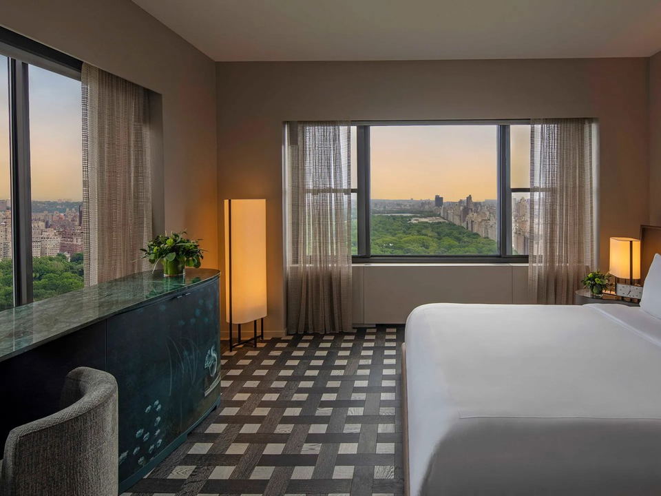 A luxury hotel room at sunset with floor-to-ceiling windows offering a panoramic view of Central Park and the Manhattan skyline.