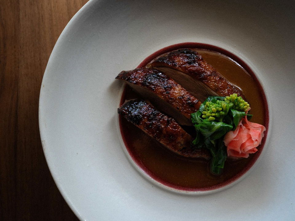 Glazed duck slices with broccolini and pickled ginger in a rich sauce, served in a white bowl.