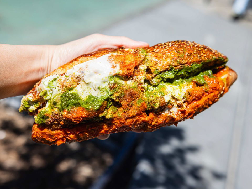A hand holds a large, generously stuffed sandwich on a sesame-seeded roll with bright green herb sauce, fried protein, and white sauce visible in