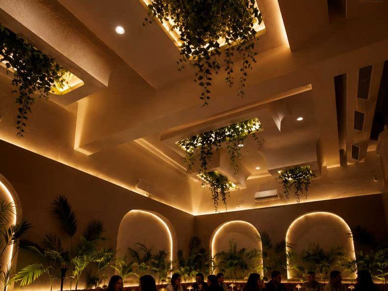 A warmly lit restaurant interior with arched LED-backlit niches, tropical palms, hanging greenery, velvet lounge seating, and candlelit tables.