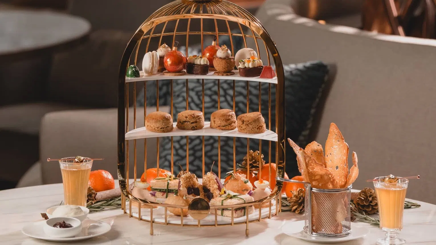 A golden birdcage-shaped stand holds a variety of pastries, scones, and sandwiches, flanked by two glasses of juice and oranges on a marble table.