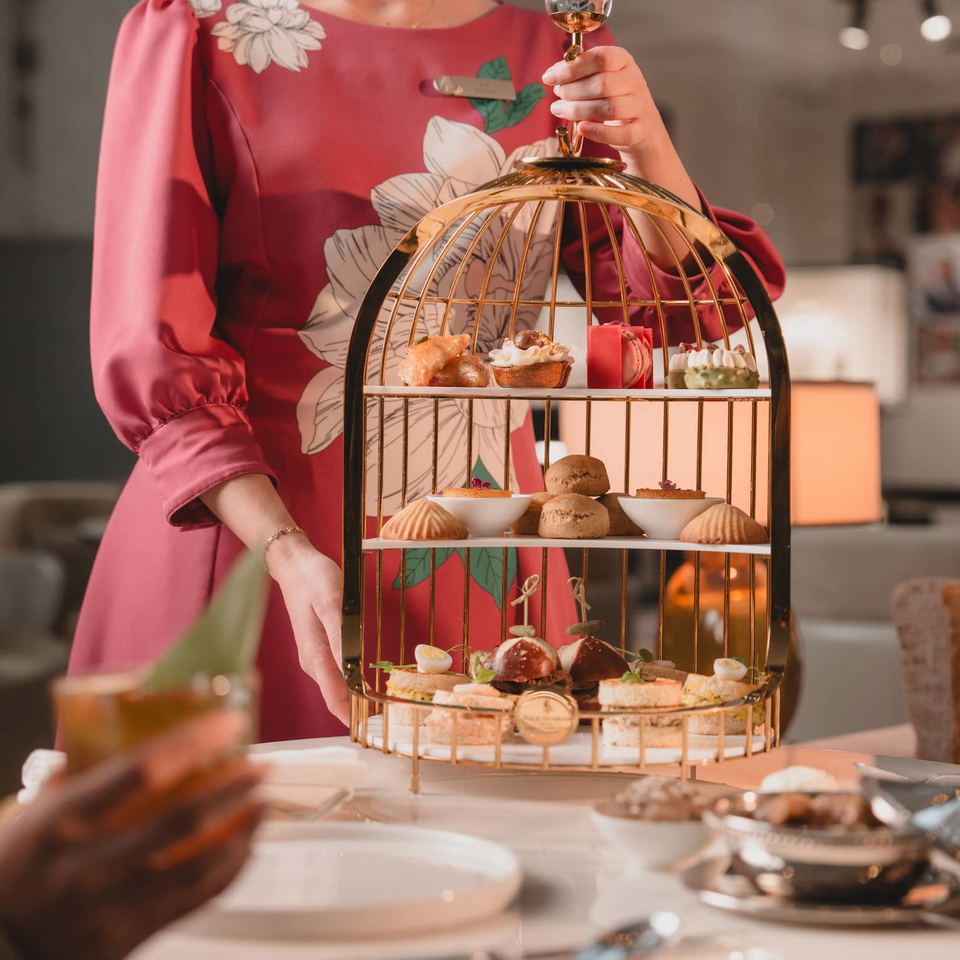 A woman in a floral dress holds a golden birdcage-shaped stand filled with various pastries and small sandwiches for high tea.