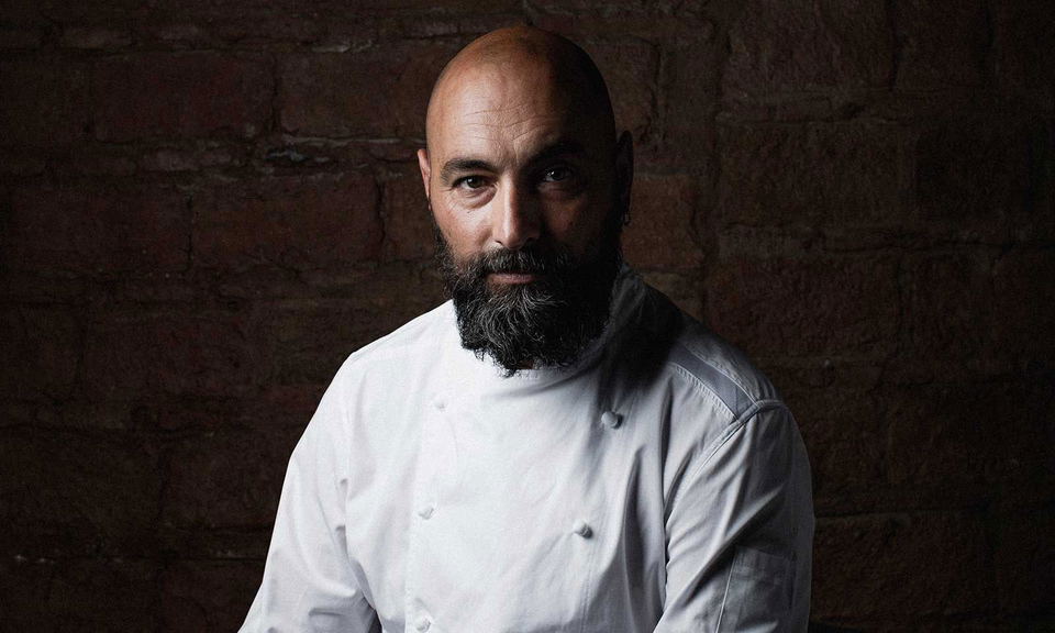 Professional portrait of a bald man with a full beard wearing a white chef's jacket against a textured brick wall