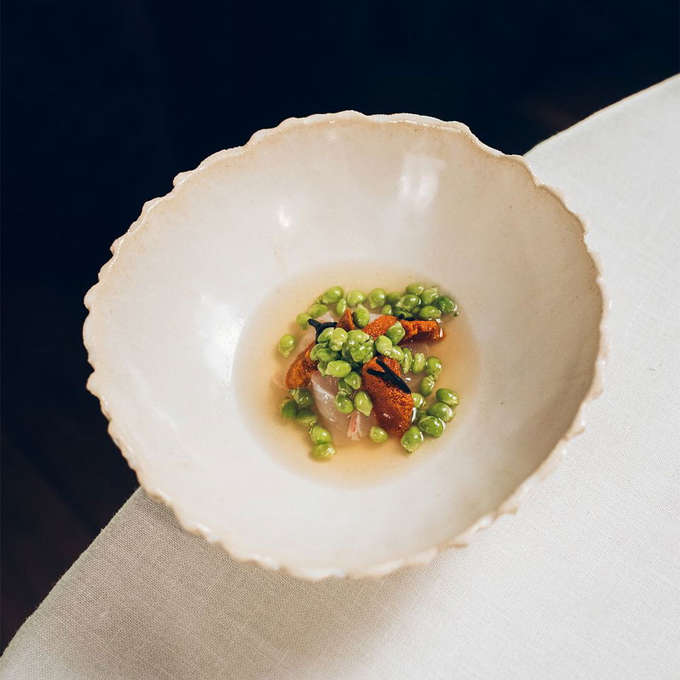 An elegant plated dish showing fire-cooked fish with bright green peas in a light broth, presented in a handcrafted ceramic bowl