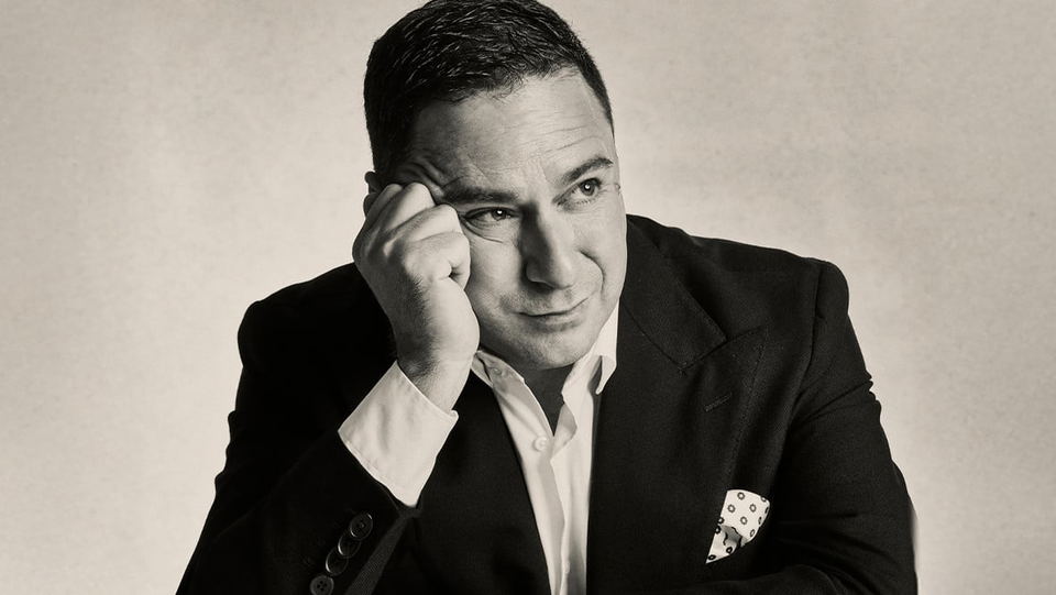 Professional black-and-white portrait of a man in a dark suit with pocket square, hand to temple in a thoughtful pose
