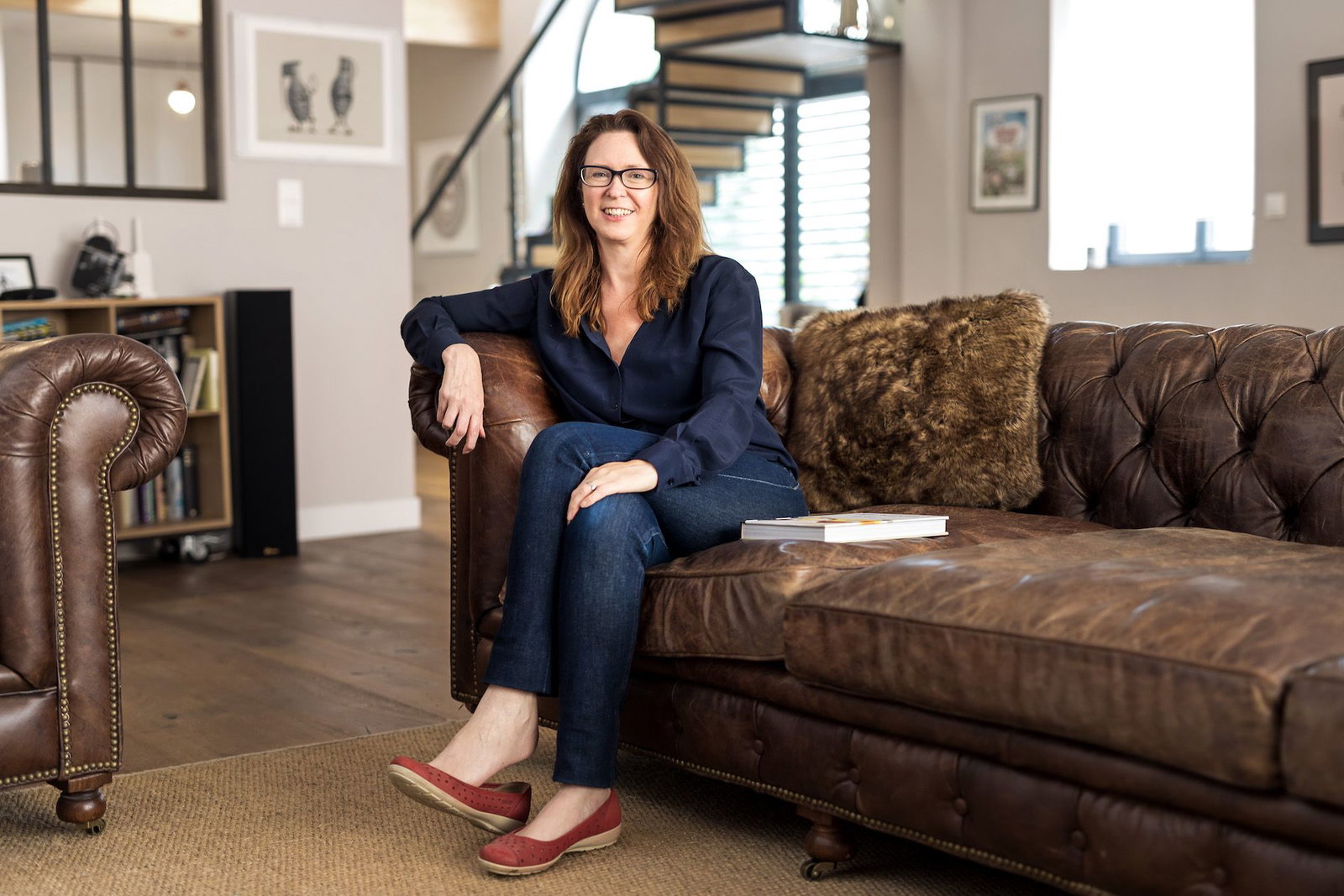 Jane Anson, a woman with long brown hair and glasses, smiles while seated on a brown leather couch in a living room.