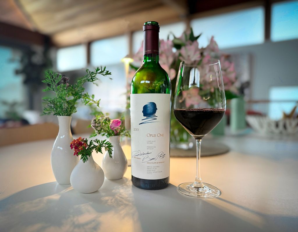 A bottle of Opus One 2013, a glass of red wine, and three small white vases with flowers are arranged on a white table.