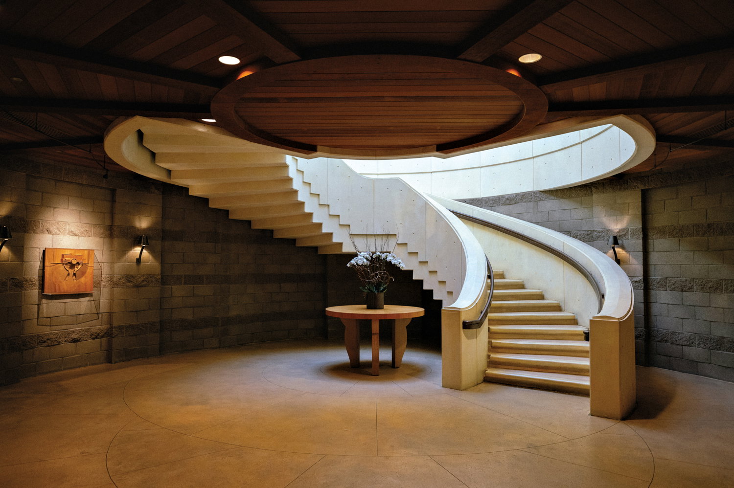 A grand, curved staircase with light-colored steps and a dark railing, illuminated by natural light from above and spotlights on the walls.