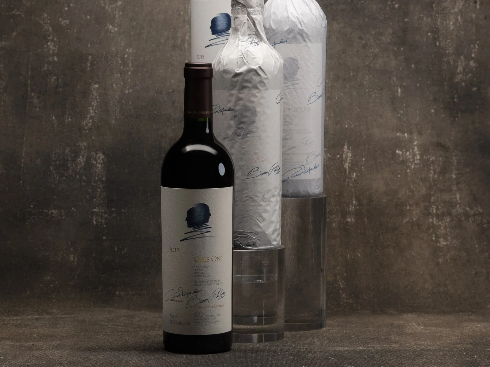Three bottles of Opus One wine, two wrapped in white paper, one unwrapped with a visible 2013 label, stand on a dark surface.