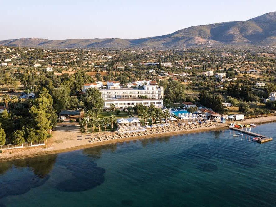 Aerial view of Paradise Resort Evia in Eretria, featuring a multi-story white hotel building, a swimming pool, a sandy beach lined with loungers and