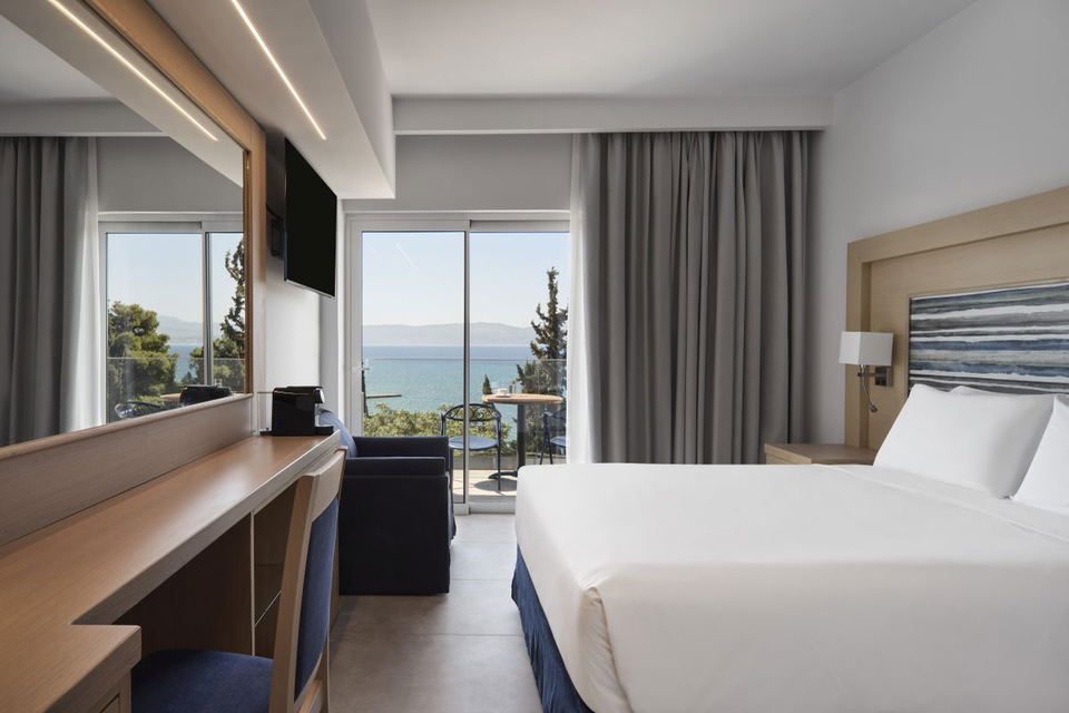 Bright hotel room at Paradise Resort Evia with a king bed, wooden desk, wall-mounted TV, and sliding glass doors opening to a balcony with sea and mountain views.