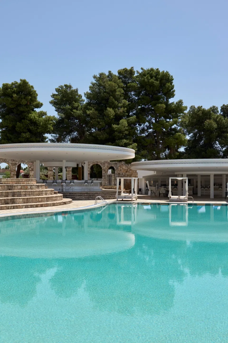 A large, inviting swimming pool with turquoise water, reflecting the surrounding white buildings and lush green trees under a clear blue sky.