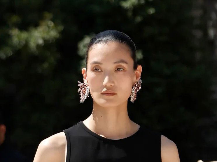 A woman with dark hair pulled back wearing a black tank top and large sparkly drop earrings, photographed outdoors with foliage in the background.